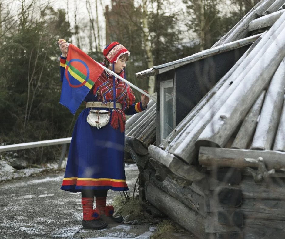 Kvinna i samisk folkdräkt hissar den samiska flaggan vid en timrad byggnad en vinterdag.