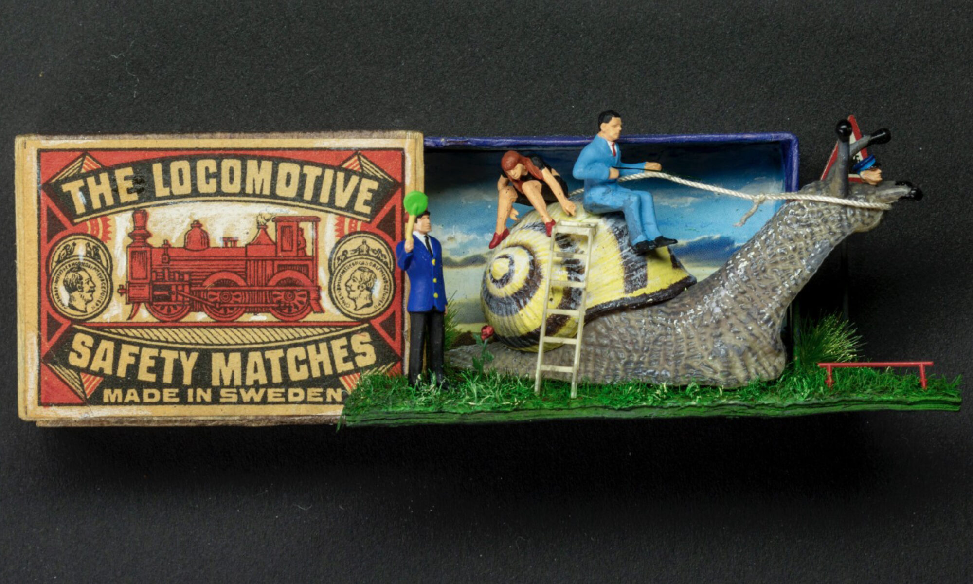 Tändsticksask med etiketten “The Locomotive Safety Matches, Made in Sweden” bredvid en liten diorama där miniatyrfigurer klättrar upp på en stor snigel, mot en mörk bakgrund.